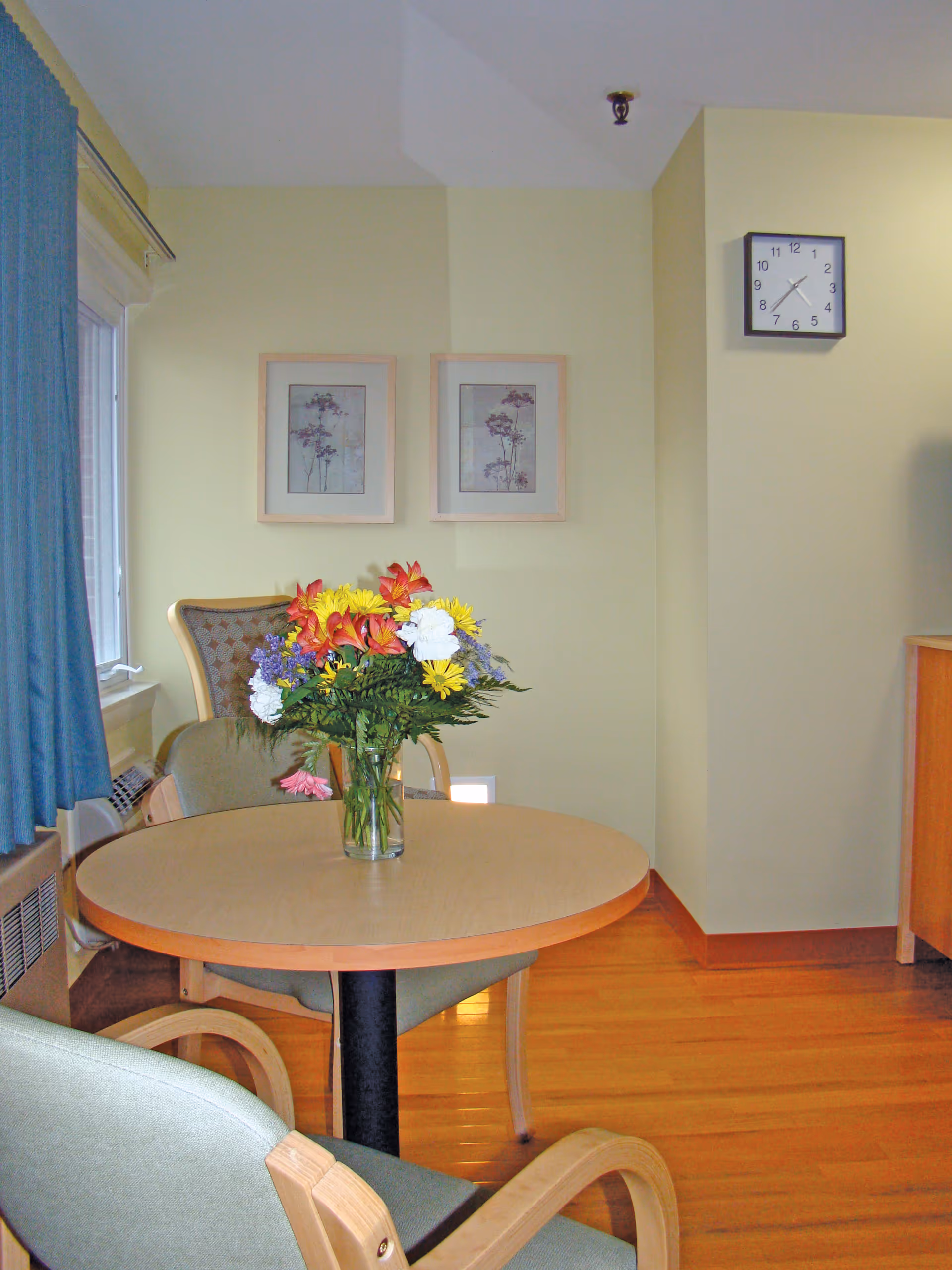 A small round wooden table with a glass vase of colorful flowers on top, surrounded by three chairs with light green cushions. The room has light yellow walls, two framed floral pictures on the wall, a square wall clock showing 11:15, a window with blue curtains, and a wooden floor.