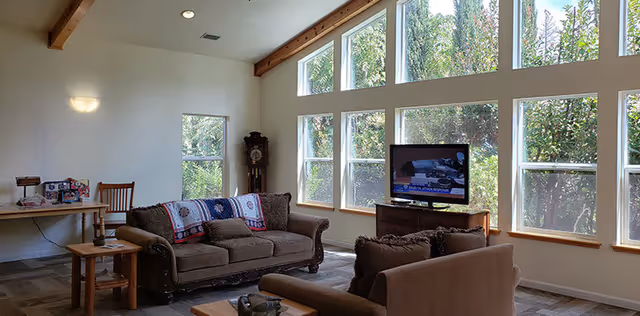 Sunlit living room with a wall of tall windows, two sofas, a coffee table, a TV on a stand and a small desk with chair.