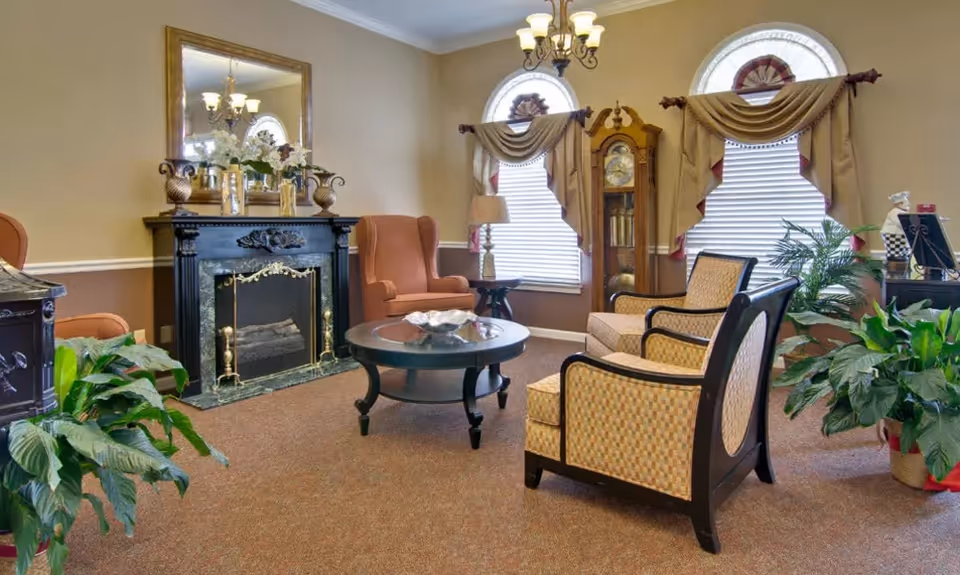 Decorative sitting area with upholstered chairs around a coffee table, a fireplace, grandfather clock, window drapes and potted plants.