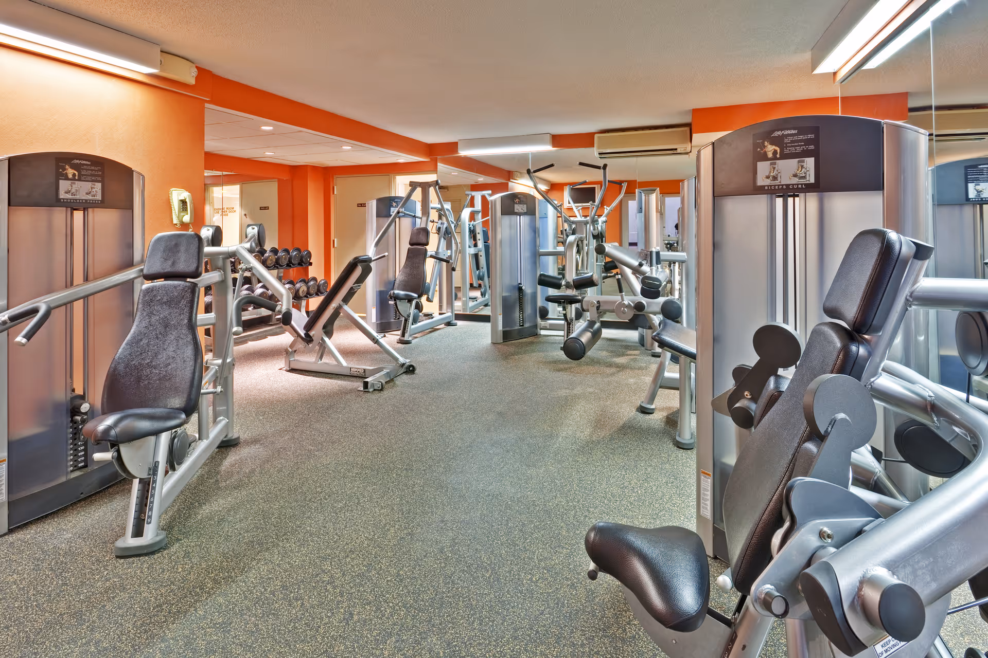 Interior view of a fitness room with various exercise machines including weight machines and dumbbells, orange walls, and mirrors along the walls.