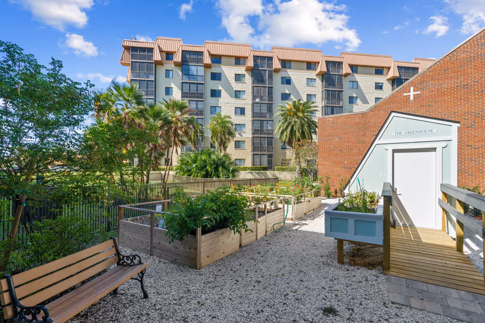 Outdoor garden area with raised wooden planter boxes filled with plants, a wooden bench on the left, and a small building labeled 'The Greenhouse' on the right. In the background, there are palm trees and a multi-story residential building under a blue sky with some clouds.
