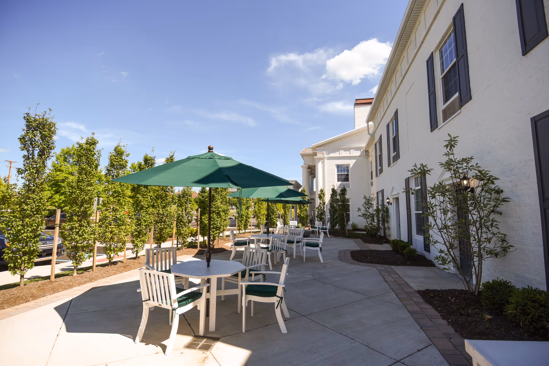 Outdoor patio area at Highgrove at Tates Creek with white tables and chairs under green umbrellas, surrounded by small trees and shrubs next to a white building with black shutters.