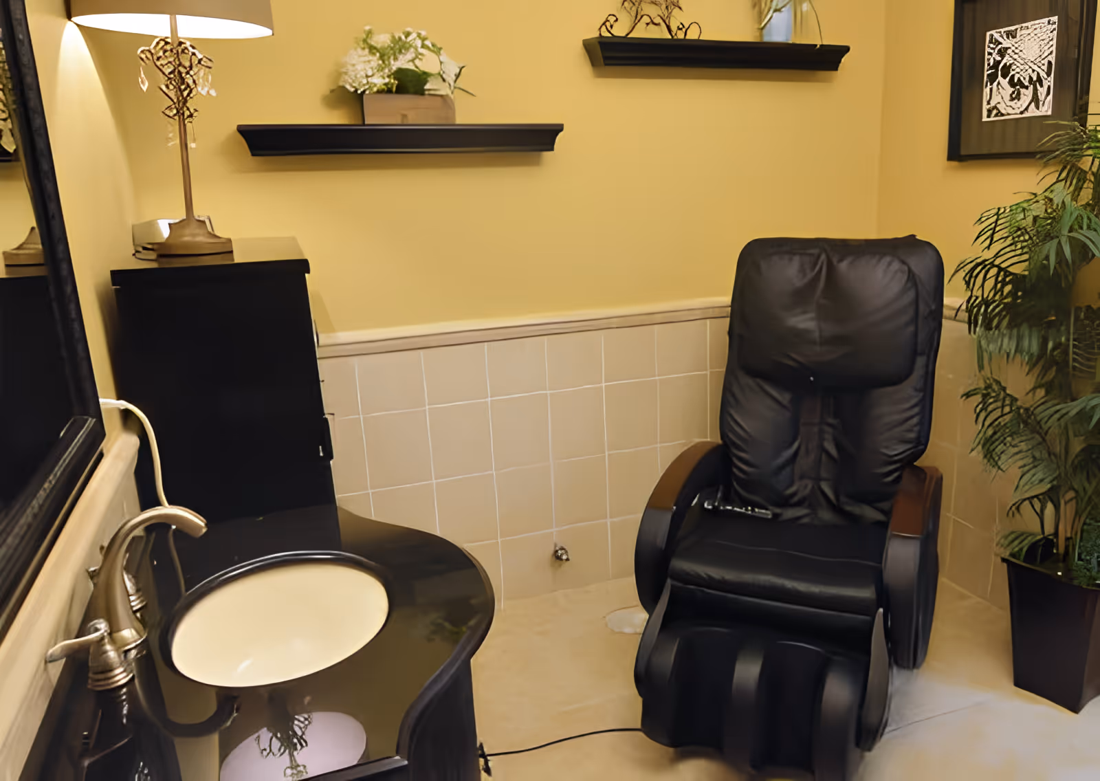 A small room with beige tiled walls and a black massage chair. There is a black vanity with a cream-colored sink and a silver faucet on the left side. A lamp with a decorative base is on the vanity, and two small shelves with decorative items are mounted on the wall. A potted plant is in the corner on the right.