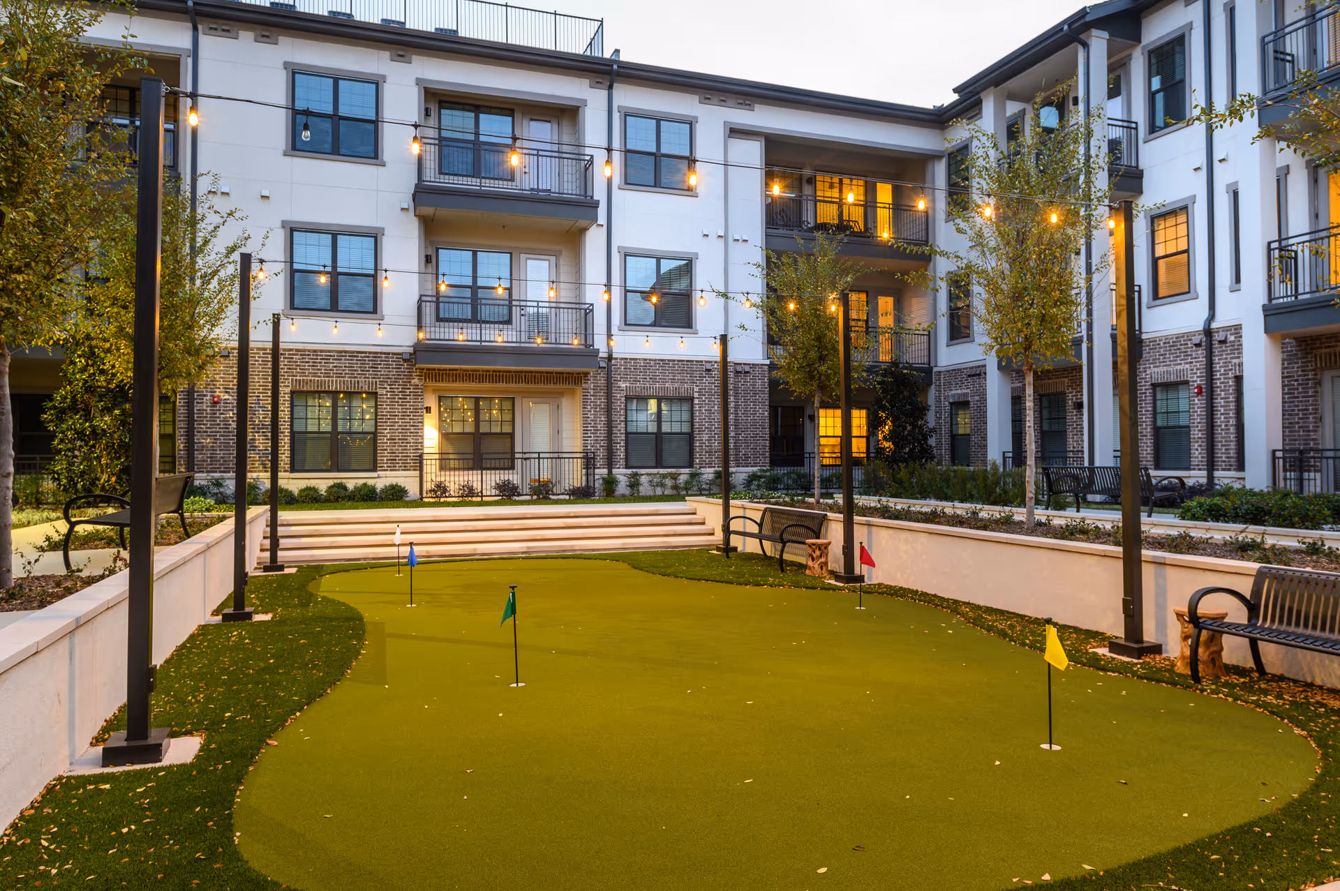 Outdoor courtyard area of Everleigh Forestwood 55+ Apartment Homes featuring a small putting green with multiple colored flags, surrounded by benches, trees, string lights, and a three-story residential building with balconies and windows in the background during dusk.
