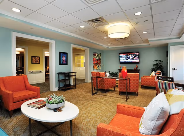A cozy living room area in a senior living facility with orange armchairs, a white oval coffee table with a plant and books, a flat-screen TV mounted on the wall, colorful artwork, and a beige patterned carpet.