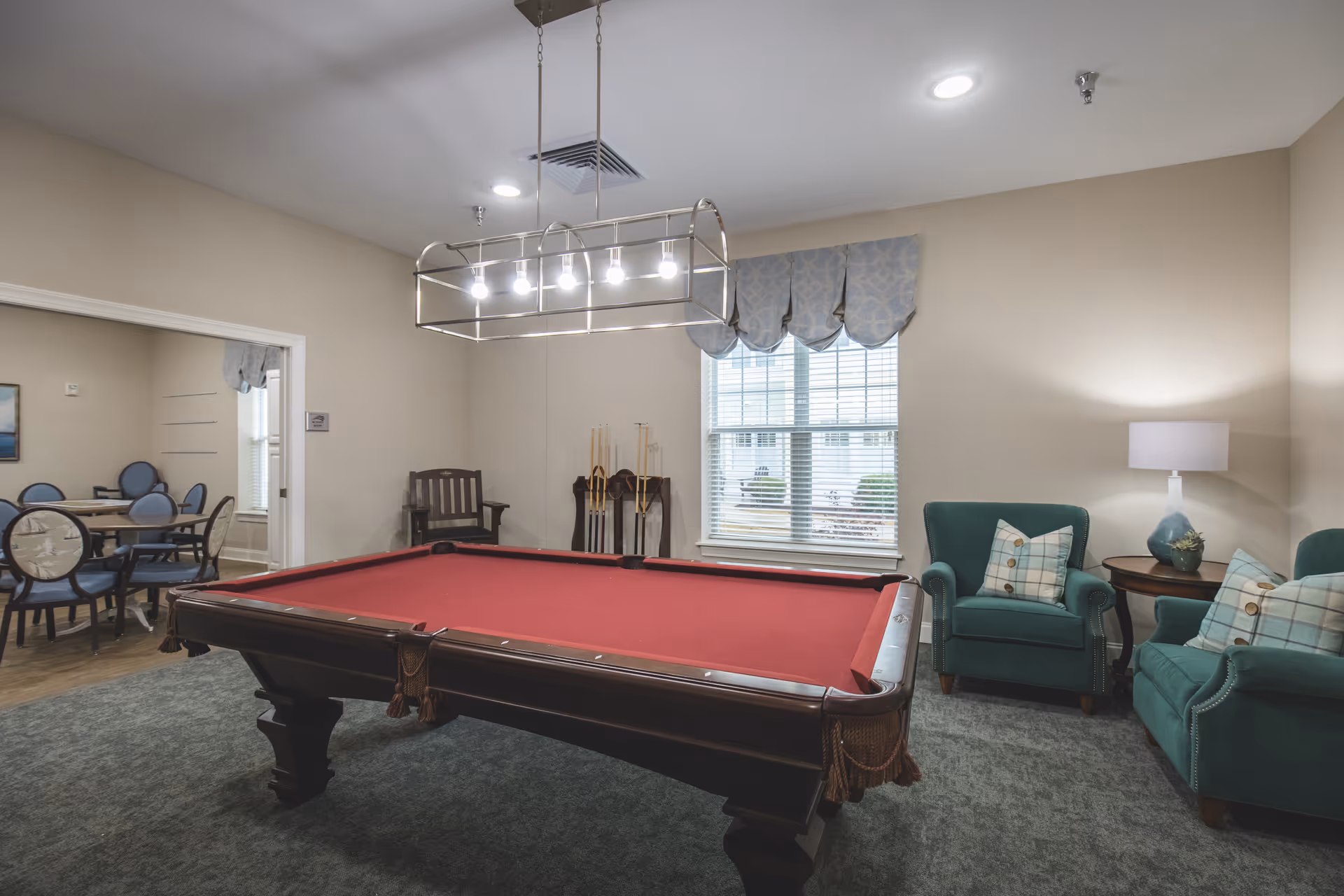 A well-lit game room featuring a red pool table in the center, two green armchairs with plaid pillows on the right, a side table with a lamp, and a window with a decorative valance. In the background, there is a dining area with round tables and chairs.