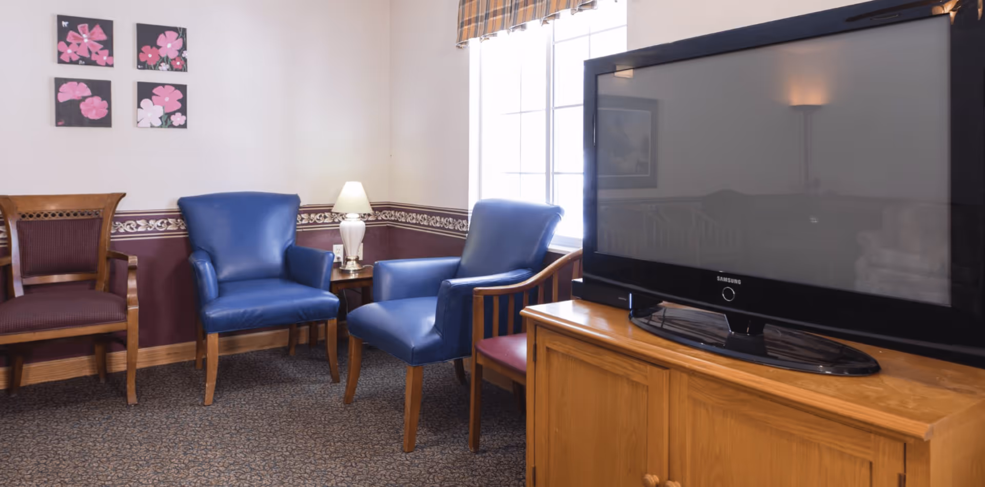 Small lounge area with blue armchairs, wooden chairs, a table lamp, and a television on a wooden cabinet by a window.