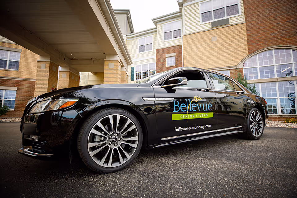 A black luxury sedan parked in front of a multi-story senior living facility building. The car has the Bellevue Senior Living logo and website printed on the side. The building features large windows and a covered entrance.