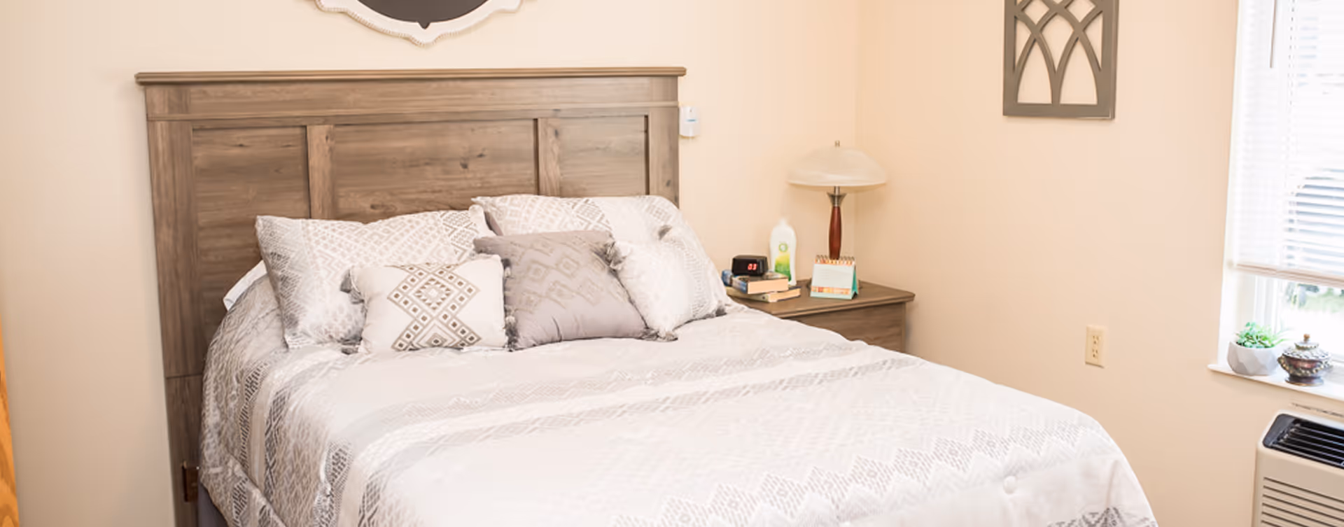 A neatly made bed with a wooden headboard, adorned with multiple patterned pillows in neutral tones. Next to the bed is a wooden nightstand with a lamp, books, a calendar, and a bottle of lotion. A window with blinds and a small plant on the windowsill is visible on the right side of the room.