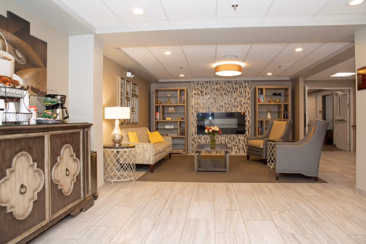 A cozy living room area in an assisted living facility featuring a patterned sofa with yellow cushions, two gray armchairs, a coffee table with a vase of flowers, built-in bookshelves, and a wall-mounted flat screen TV with a mosaic tile backdrop. To the left, there is a wooden cabinet with a coffee station on top. The room has tiled flooring and recessed lighting in the ceiling.