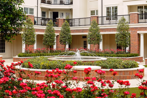 A courtyard garden with a circular fountain in the center surrounded by red flowers and neatly trimmed bushes. The courtyard is enclosed by a building with balconies and brick columns, and several small trees are planted around the fountain area.