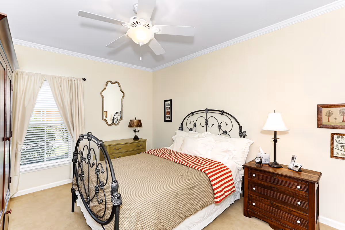 A cozy bedroom with a black metal bed frame, beige bedding, and a red and white striped throw blanket. There is a wooden dresser with a lamp, clock, and phone on the right side of the bed. On the left side, there is a green dresser with a decorative lamp and a mirror above it. A window with cream-colored curtains is on the left wall, and the room has beige walls and carpet with a white ceiling fan overhead.
