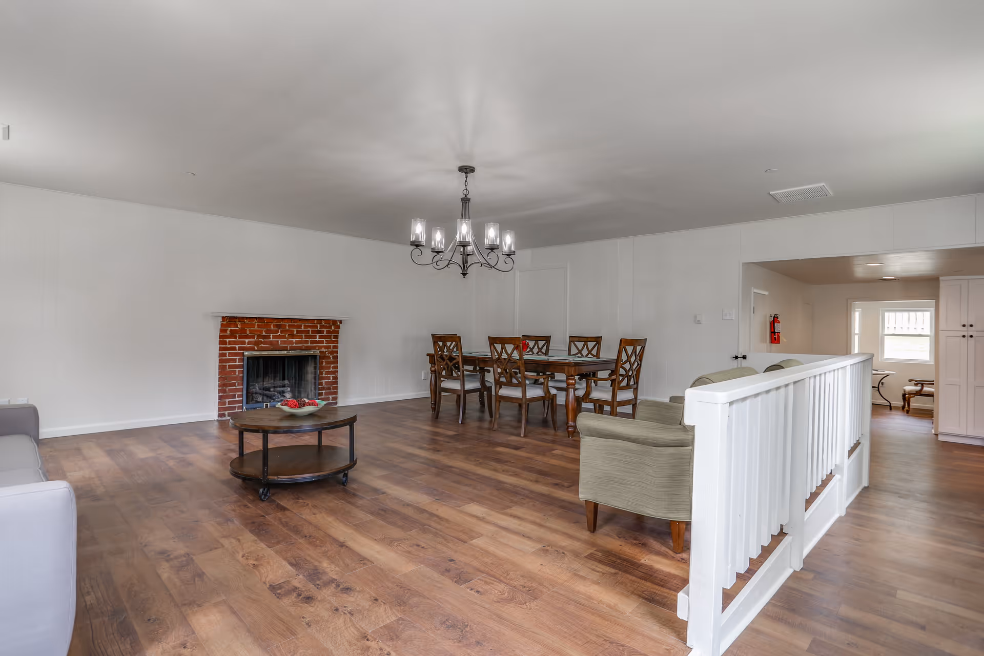 A spacious living and dining area with wooden flooring, a brick fireplace, a round coffee table, a dining table with six chairs, and a chandelier hanging from the ceiling. The walls are white, and there is a white railing separating the living area from a hallway.