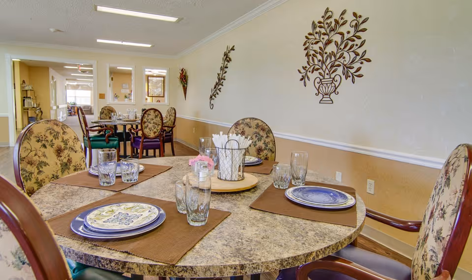 Round dining table set with plates, glasses and floral-upholstered chairs in a decorated dining room.