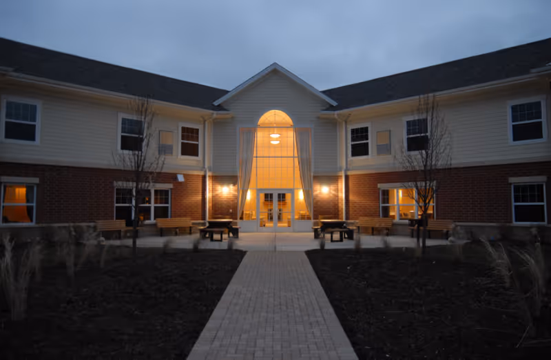 Two-story beige and brick senior living facility exterior with illuminated glass entrance.