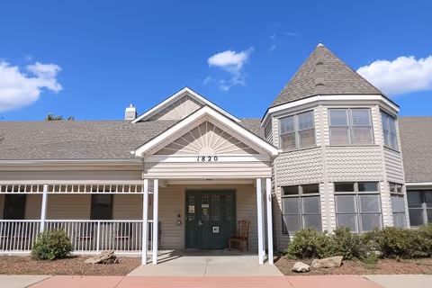Exterior front view of The Mapleton Eureka facility showing a beige building with a peaked roof, a covered entrance with the number 1820 above the door, and a turret-like structure with multiple windows on the right side. There are bushes and rocks in front of the building and a clear blue sky with a few clouds above.