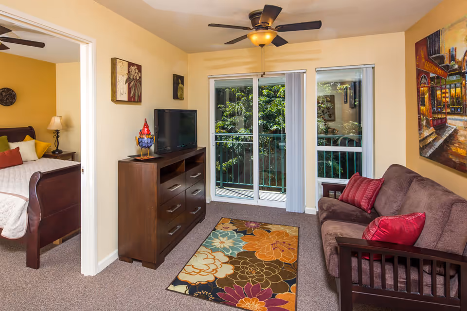 A cozy living room with a brown couch adorned with red pillows, a colorful floral rug on the carpeted floor, a wooden dresser with a flat-screen TV and a decorative rooster figurine on top. Sliding glass doors lead to a balcony with green trees visible outside. To the left, an open doorway reveals a bedroom with a wooden bed, beige bedding, and a bedside table with a lamp.