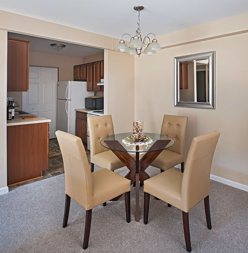 A dining area with a round glass table and four beige upholstered chairs on a carpeted floor. A decorative centerpiece is on the table. Behind the dining area is a kitchen with wooden cabinets, a white refrigerator, a microwave, and a stove. A silver-framed mirror hangs on the wall, and a chandelier with five lights is above the table.