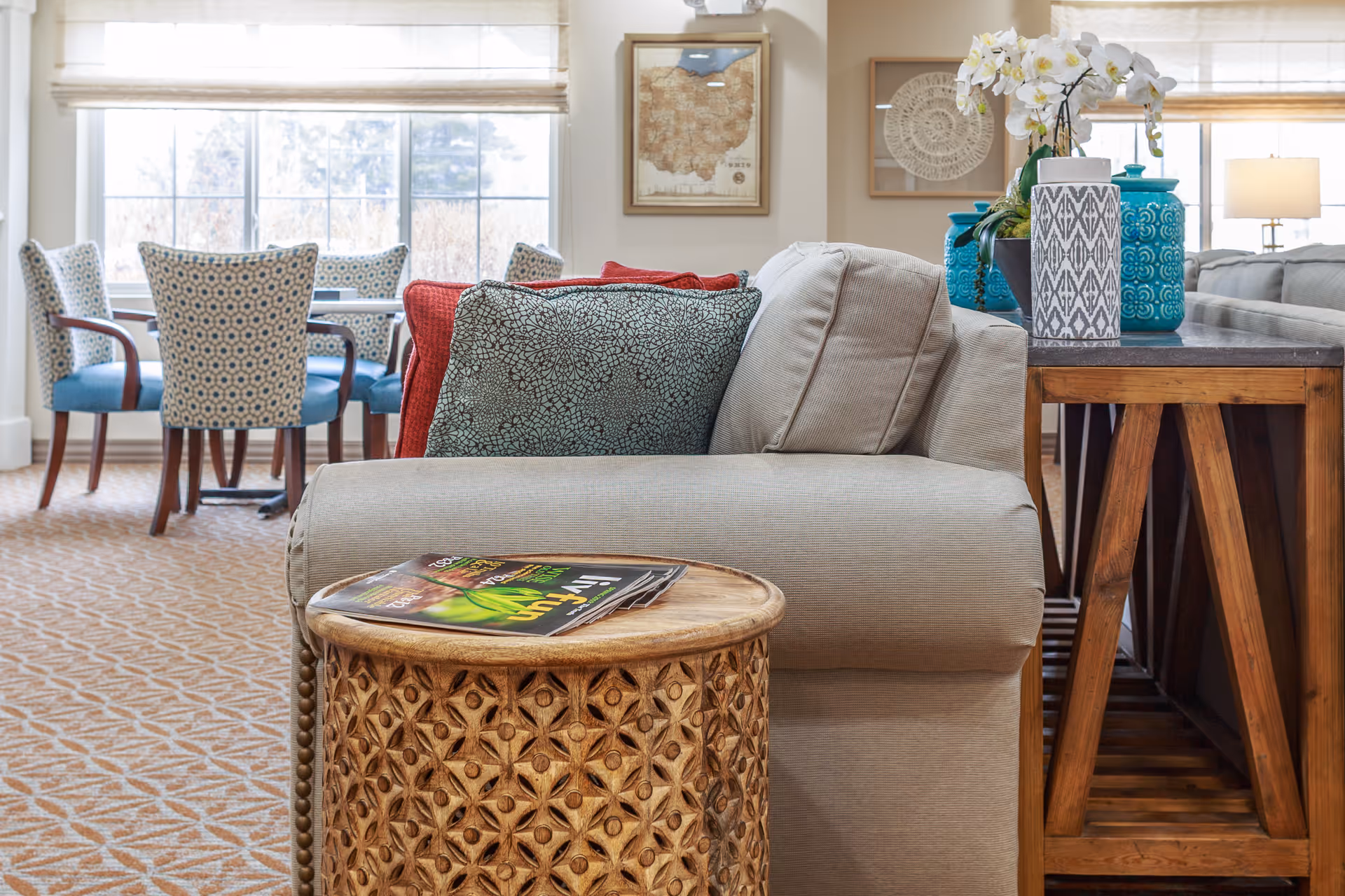 A cozy living room area with a beige sofa adorned with patterned cushions in red and blue. In front of the sofa is a round wooden side table with a carved floral design, holding a magazine. Behind the sofa is a wooden console table with decorative blue vases and a white orchid plant. In the background, there is a dining area with a table and four upholstered chairs with a blue and white pattern, positioned in front of large windows letting in natural light. The walls have framed artwork, and the floor is carpeted with a geometric pattern.
