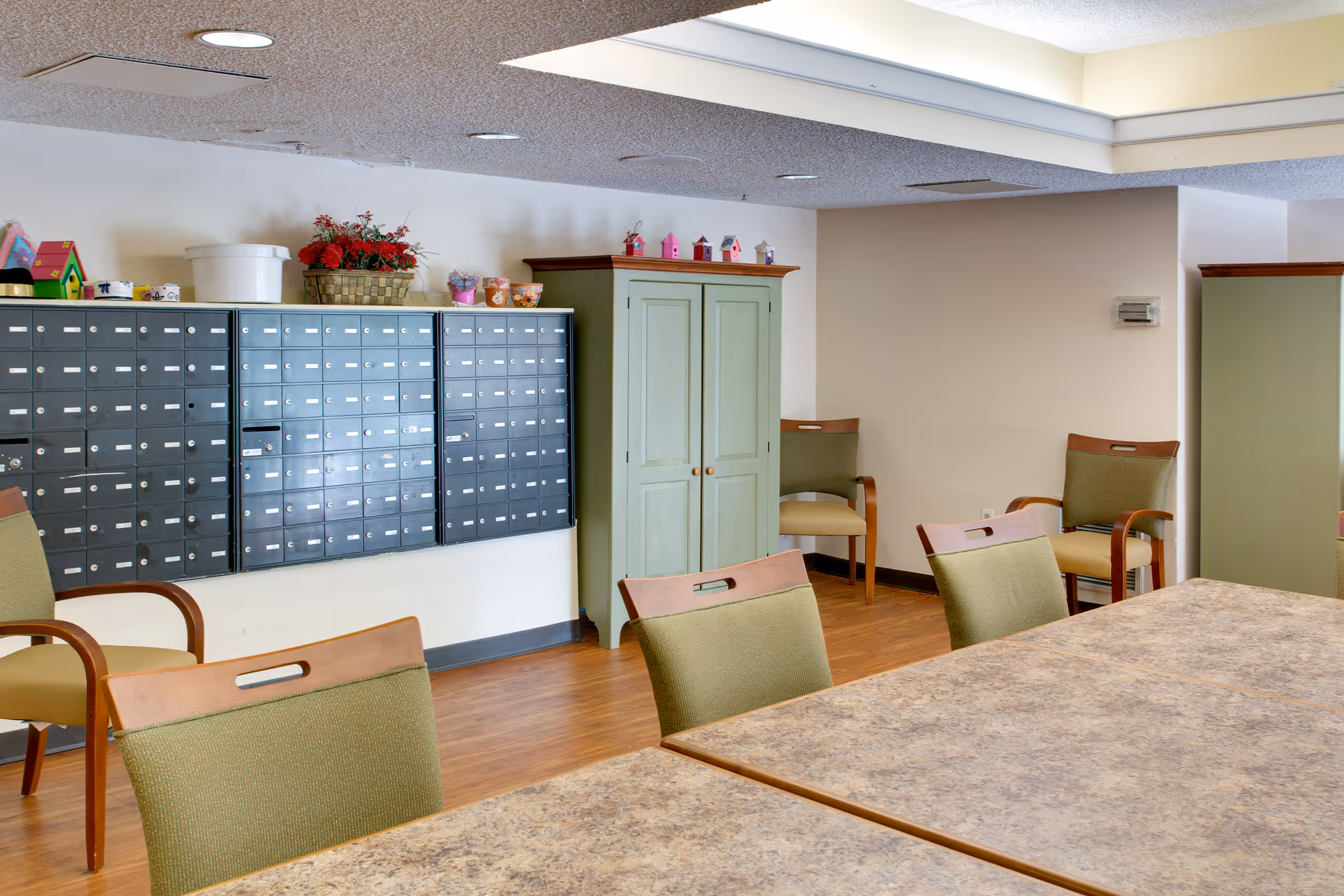 Interior room with a set of green cushioned chairs around a large table with a marble-like surface. On the left wall, there are multiple black mailboxes mounted, with decorative items such as small birdhouses and a basket with red flowers placed on top of a green cabinet. The room has wooden flooring and a white ceiling with recessed lighting.