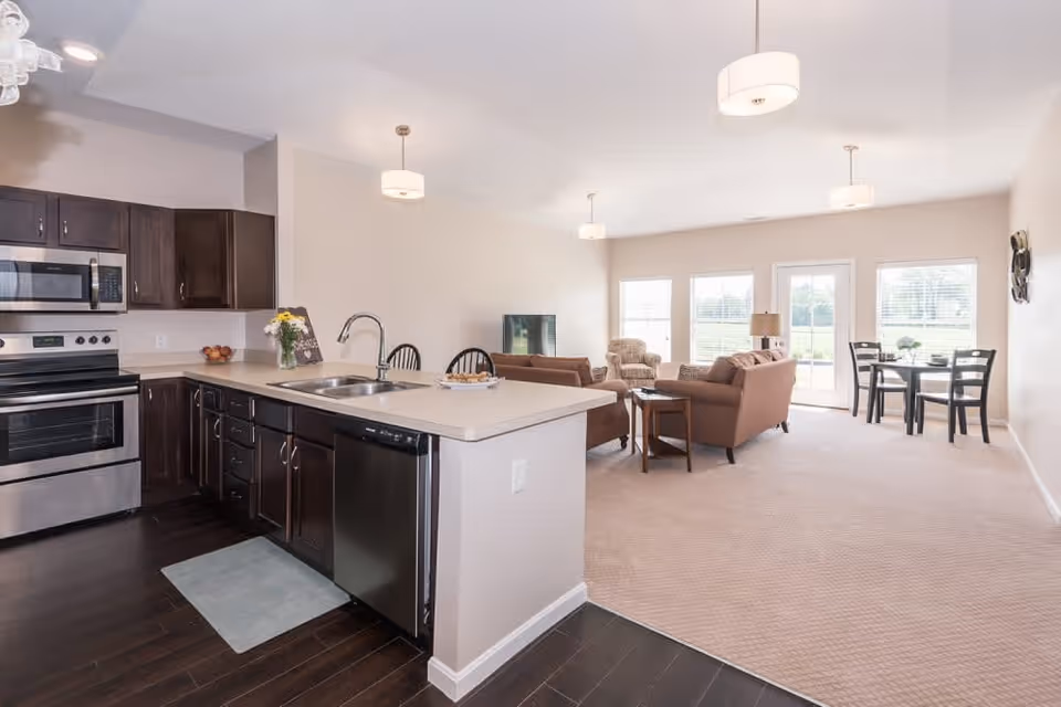 Open concept interior of a senior living facility showing a kitchen with dark wood cabinets, stainless steel appliances, and a sink island. Adjacent to the kitchen is a living area with brown sofas, a TV, a small side table, and a dining area with a round table and four chairs near large windows and a glass door letting in natural light.