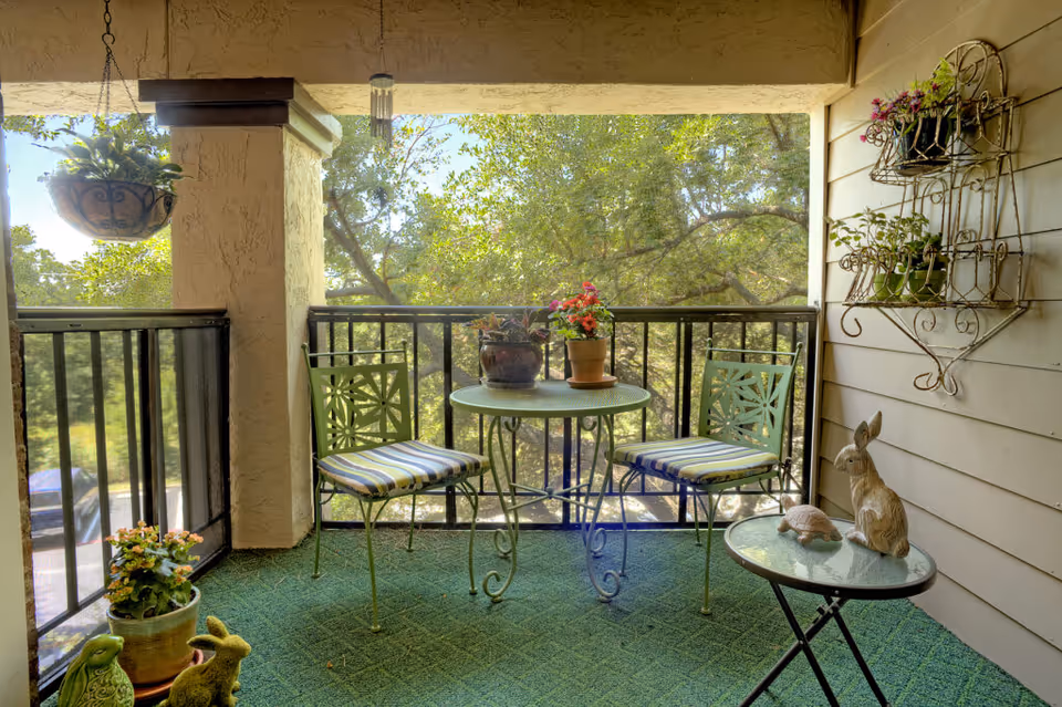A cozy balcony with green seating, a small round table, and potted plants, surrounded by trees.