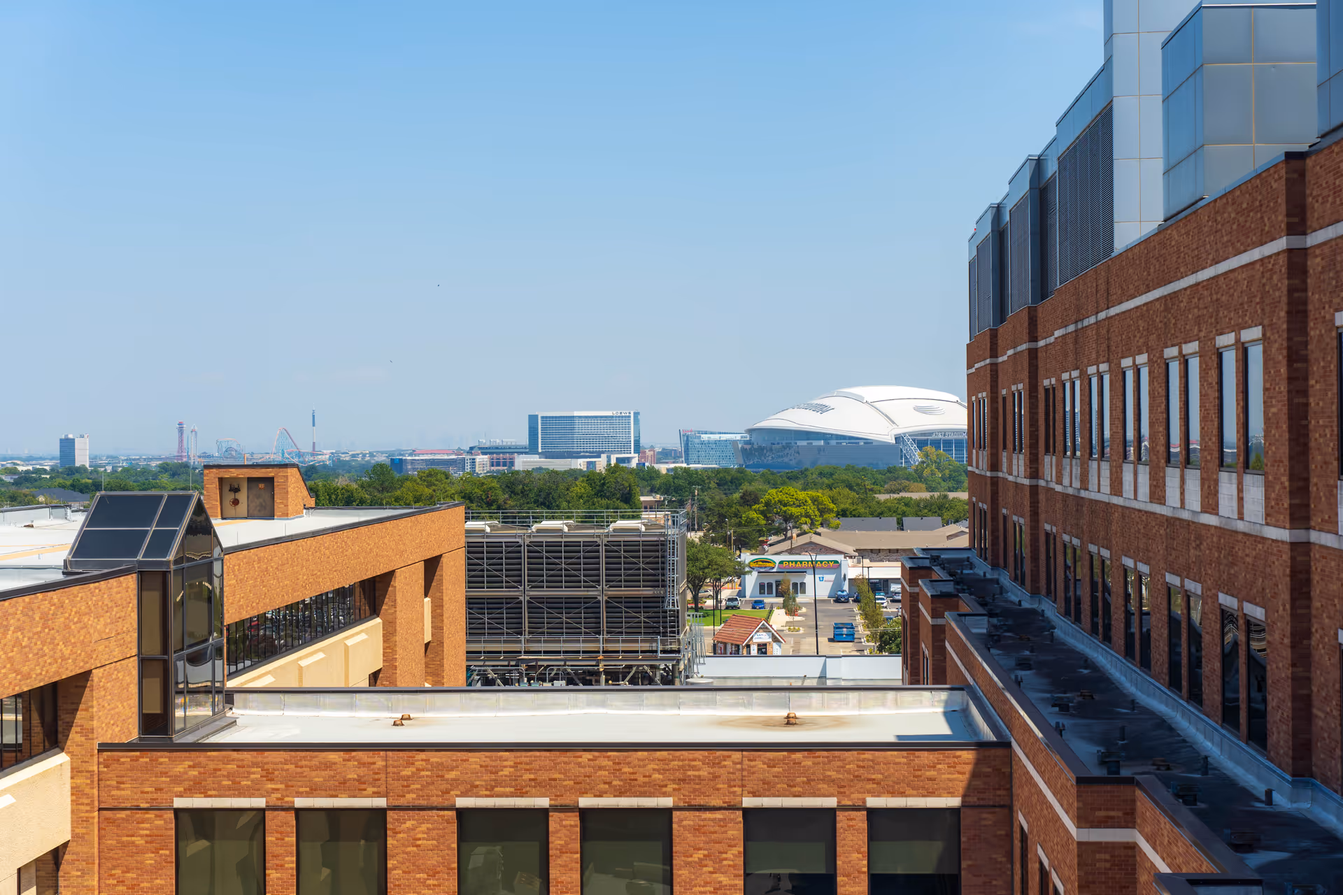 Rooftop view across adjacent brick hospital/office buildings toward a distant white-domed stadium and city skyline under a clear blue sky.