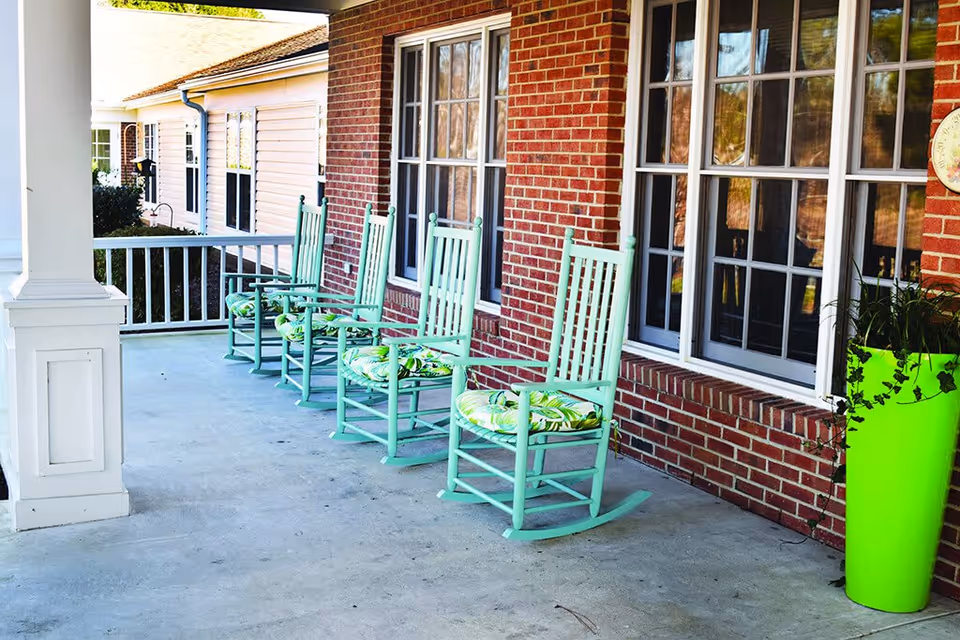 A covered outdoor porch area with four light green rocking chairs with patterned cushions lined up against a red brick wall with large windows. A bright green planter with trailing plants is placed near the chairs.