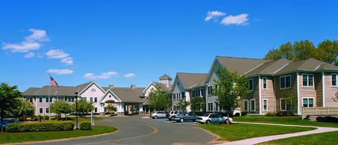 Exterior view of a senior living facility with multiple connected buildings, a circular driveway with parked cars, well-maintained landscaping, and a clear blue sky with a few clouds.