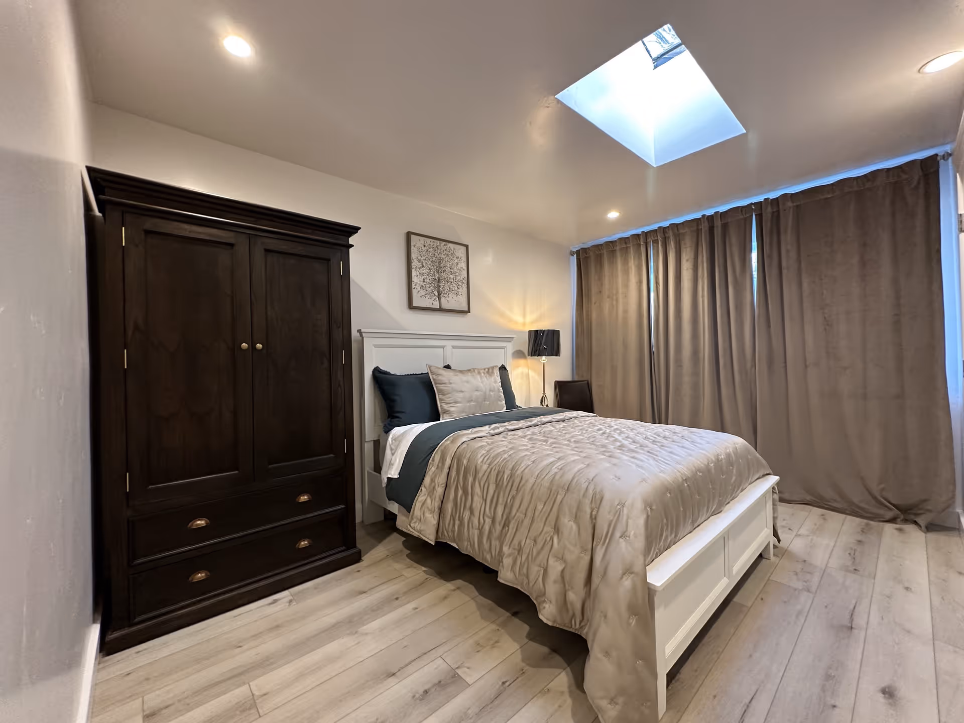 A cozy bedroom with a white wooden bed frame, beige comforter, and dark blue pillows. There is a dark wooden wardrobe to the left of the bed, a small lamp on a nightstand, and a framed picture on the wall above the bed. The room has light wood flooring, beige curtains covering the window, and a skylight on the ceiling letting in natural light.
