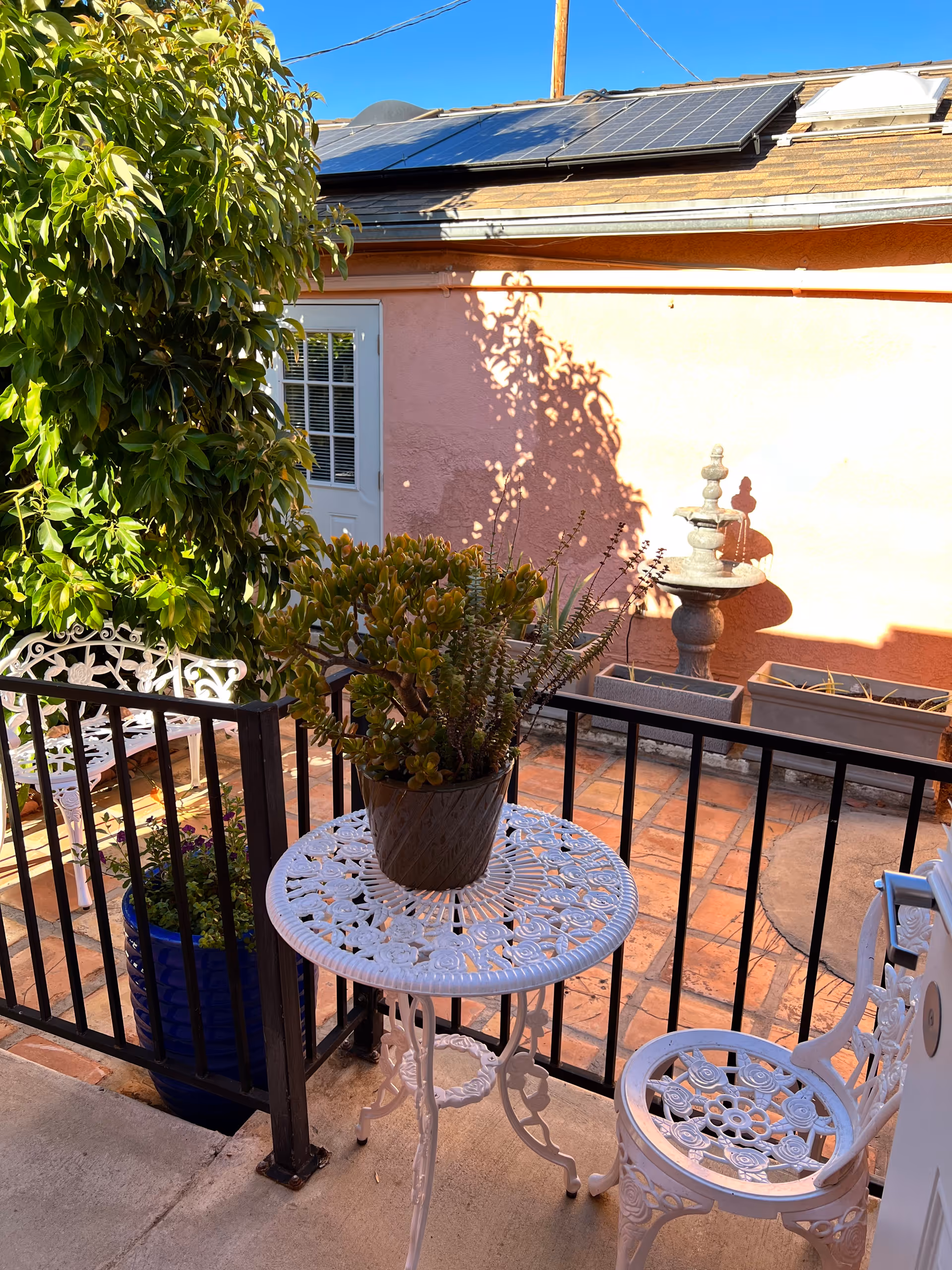 A small outdoor patio area with a white ornate metal table and chair. A potted plant sits on the table. There is a black metal railing around the patio, a large leafy plant in a blue pot, and a pink wall with a white door and a small fountain in the background. Solar panels are visible on the roof above the wall.