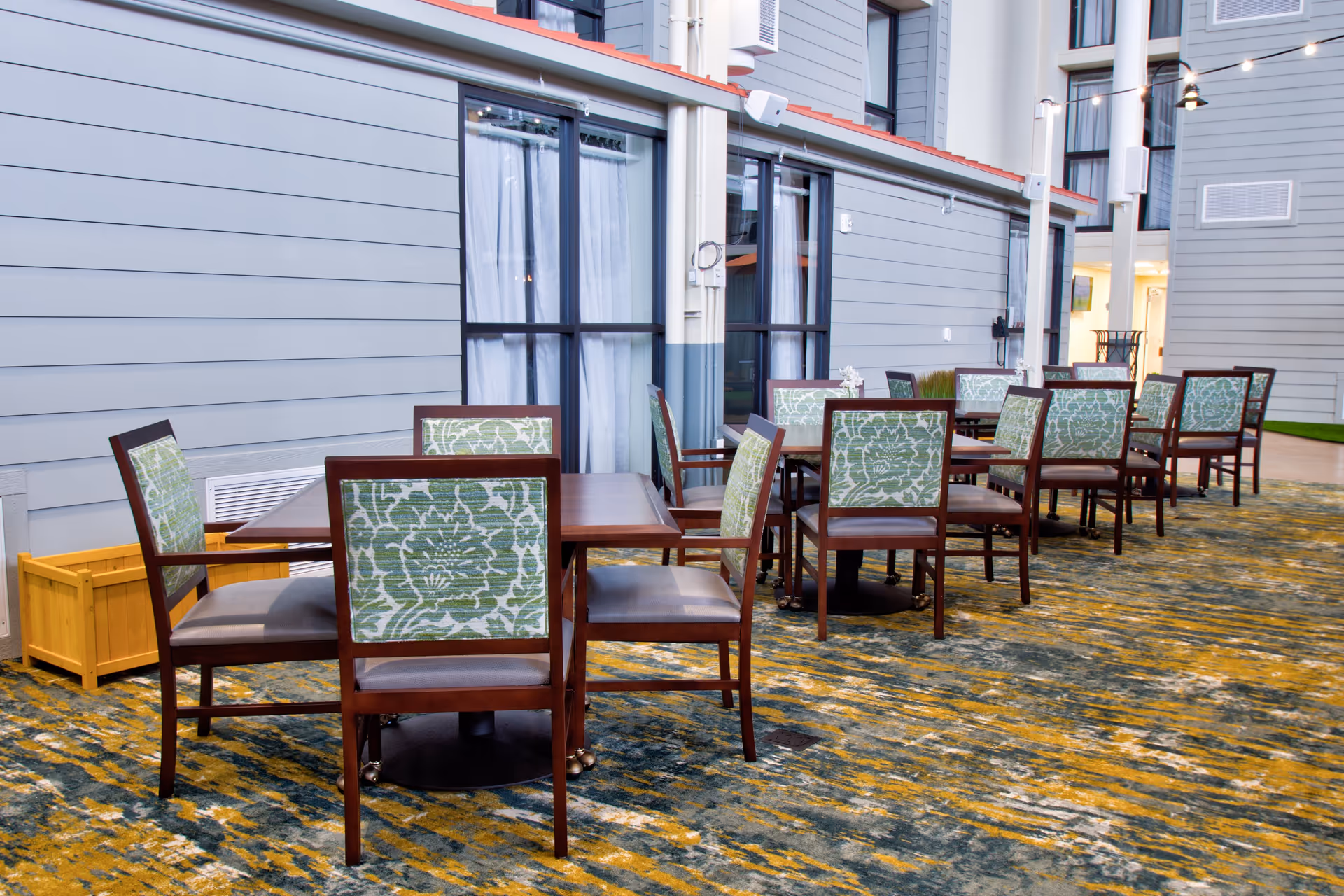 Indoor seating area with multiple wooden tables and chairs featuring green floral patterned backs, set against a backdrop of large windows with white curtains and gray paneled walls.