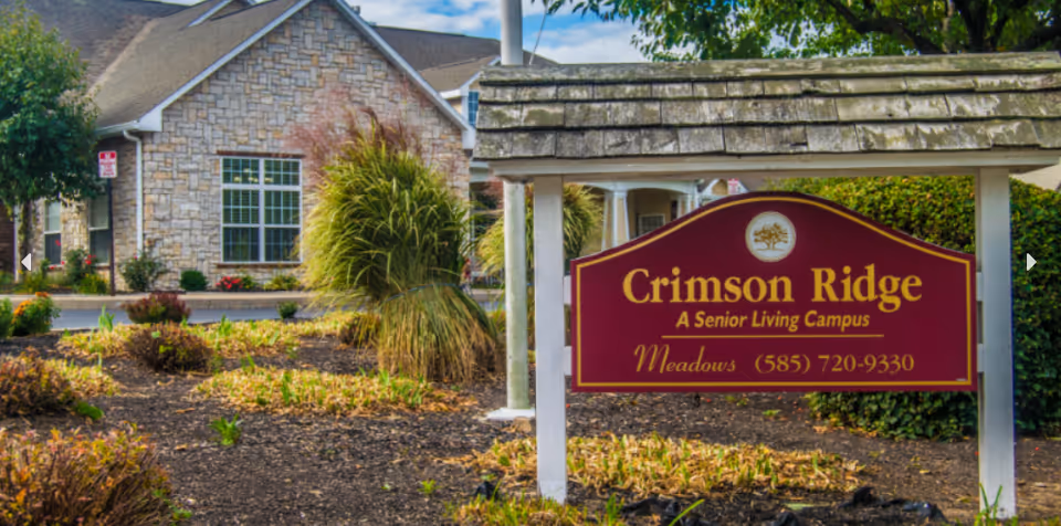 Wooden entrance sign reading 'Crimson Ridge A Senior Living Campus' in landscaped grounds with the facility building behind it.