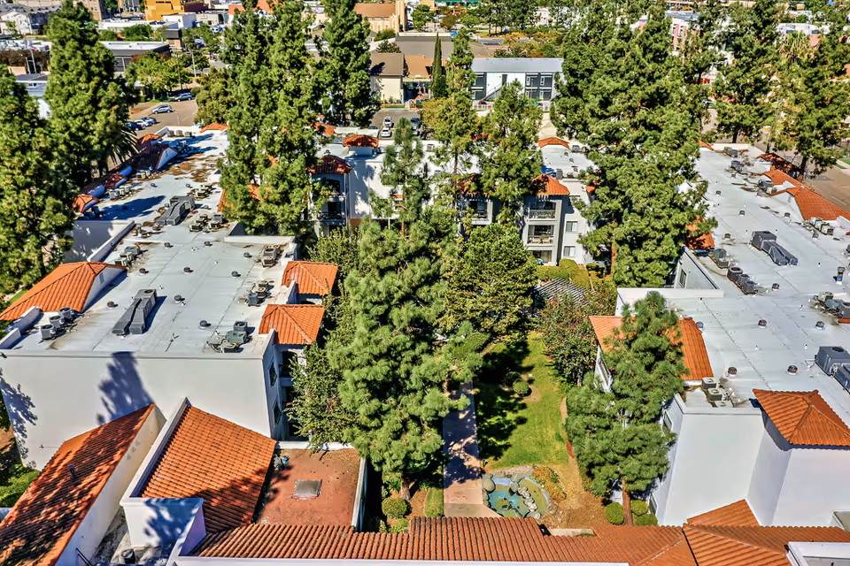 Aerial view of Solstice Senior Living at El Cajon showing white buildings with red tile roofs surrounding a green courtyard with trees and a small pond. The surrounding neighborhood and streets are visible in the background.