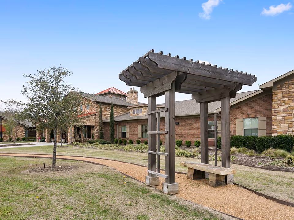 Outdoor garden area at River Oaks facility featuring a wooden pergola with a bench underneath, a curved gravel pathway, a small tree, and a brick building with stone accents in the background under a clear blue sky.