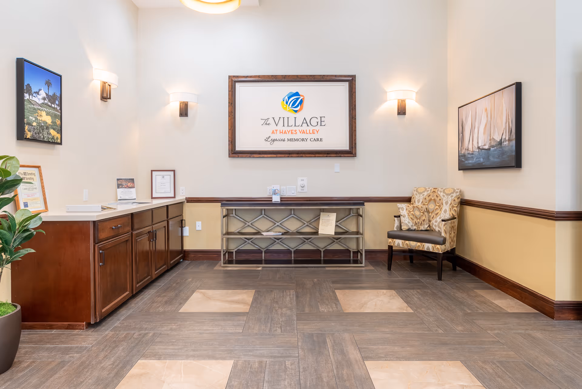 Reception-style interior with a framed 'The Village at Hayes Valley' sign above a console, cabinets to the left, and an upholstered chair to the right.