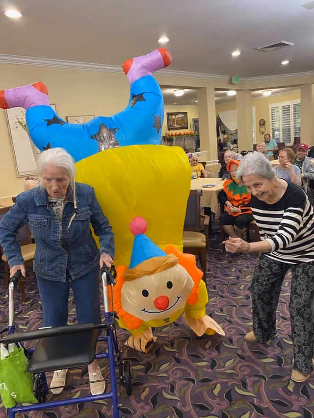 An elderly woman using a walker stands next to a person in a colorful clown costume doing a handstand in a senior living facility common area. Another elderly woman in a striped shirt gestures towards the clown. Several other seniors are seated at tables in the background, some dressed in festive clothing.
