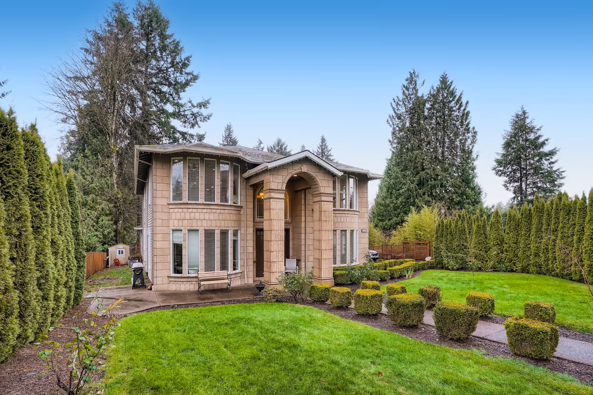 Two-story residential building with beige exterior and large windows, surrounded by a well-maintained green lawn and neatly trimmed bushes, with tall trees and a clear blue sky in the background.