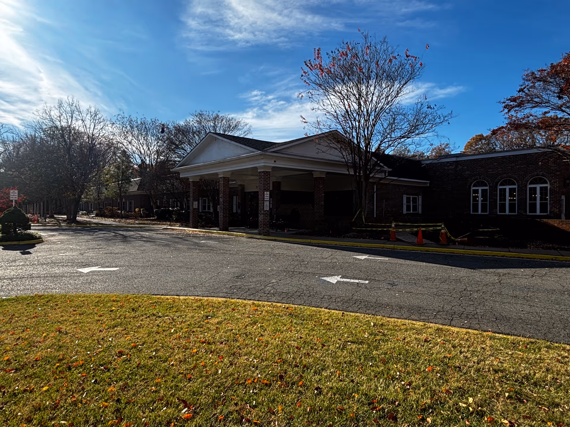 Front entrance with a covered porte-cochère at a brick building, a driveway with arrows, lawn in the foreground and trees under a blue sky.