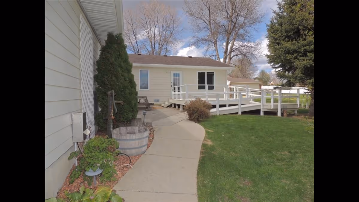 Outdoor view of a single-story building with a white wooden ramp leading to the entrance. There is a concrete pathway curving through a green lawn, with some bushes and a tree near the building. The sky is partly cloudy.