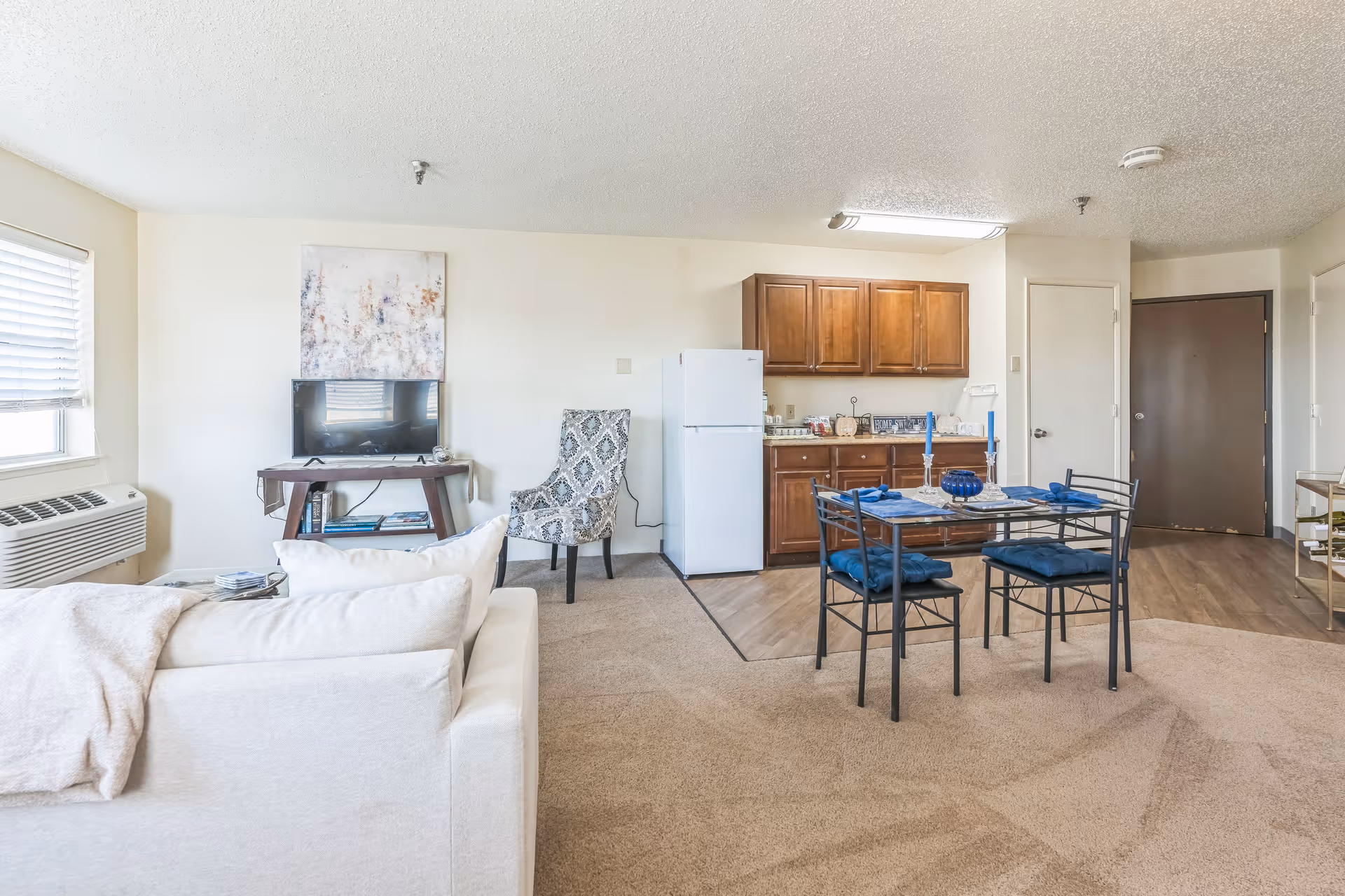 A bright and clean living area in a senior living facility featuring a white sofa with a beige throw blanket, a small TV on a wooden stand, a patterned armchair, a dining table set with blue cushions and candles, and a kitchenette with wooden cabinets and a white refrigerator. The room has beige carpet and light-colored walls with a window on the left side.