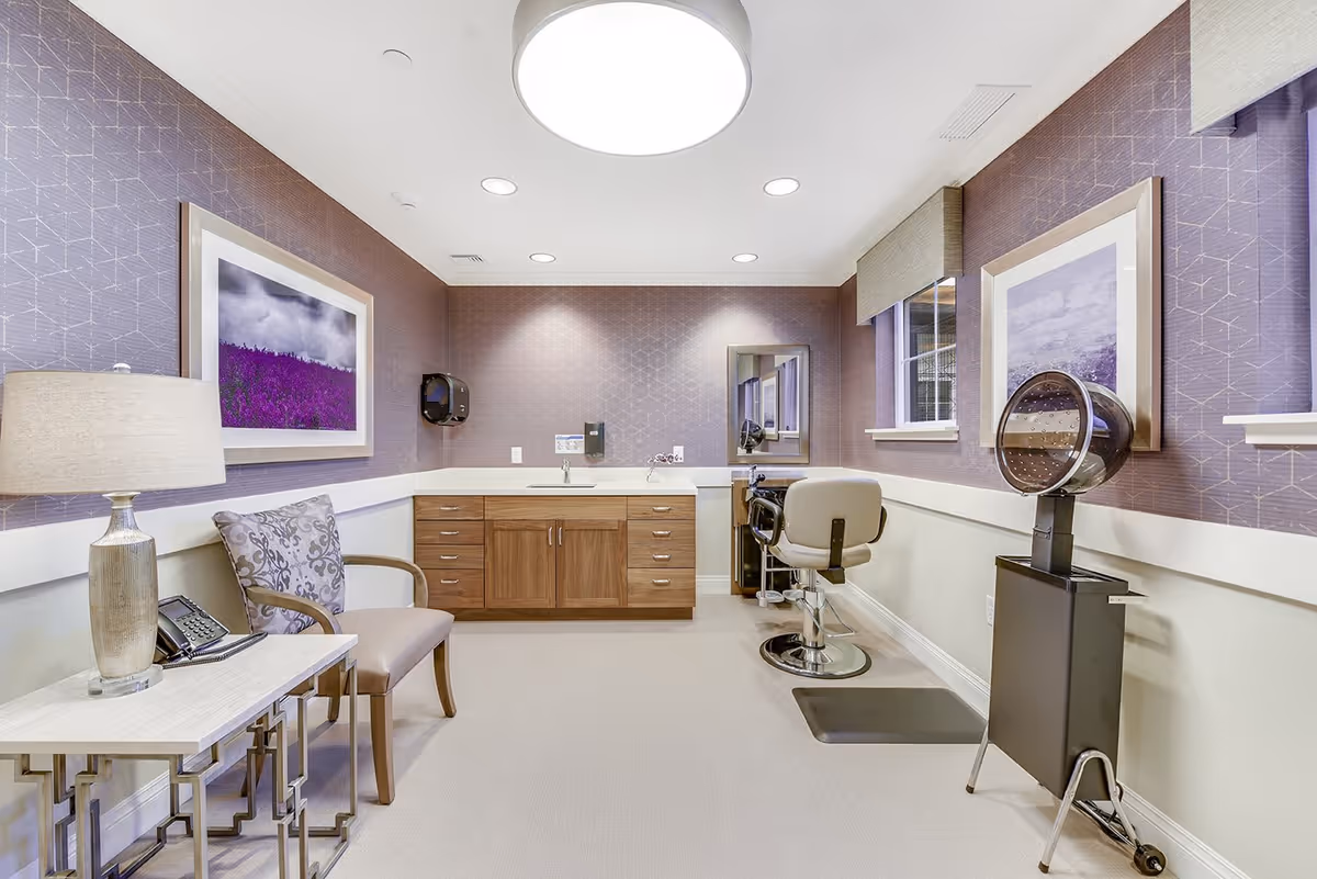 Interior room with a hair salon setup including a salon chair, hair dryer, and a mirror. The room has purple patterned wallpaper, a wooden cabinet with a sink, a chair with a patterned cushion, a table with a lamp and a phone, and framed artwork on the walls.