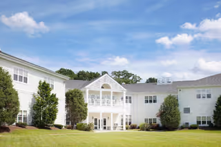 White two-story senior living building with a central covered entrance and balcony set behind a manicured lawn under a blue sky.