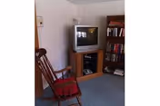 A small living room area with a wooden rocking chair with a red cushion, a wooden TV stand holding an older-style CRT television, and a bookshelf filled with books. The room has light-colored walls and blue carpeted flooring.
