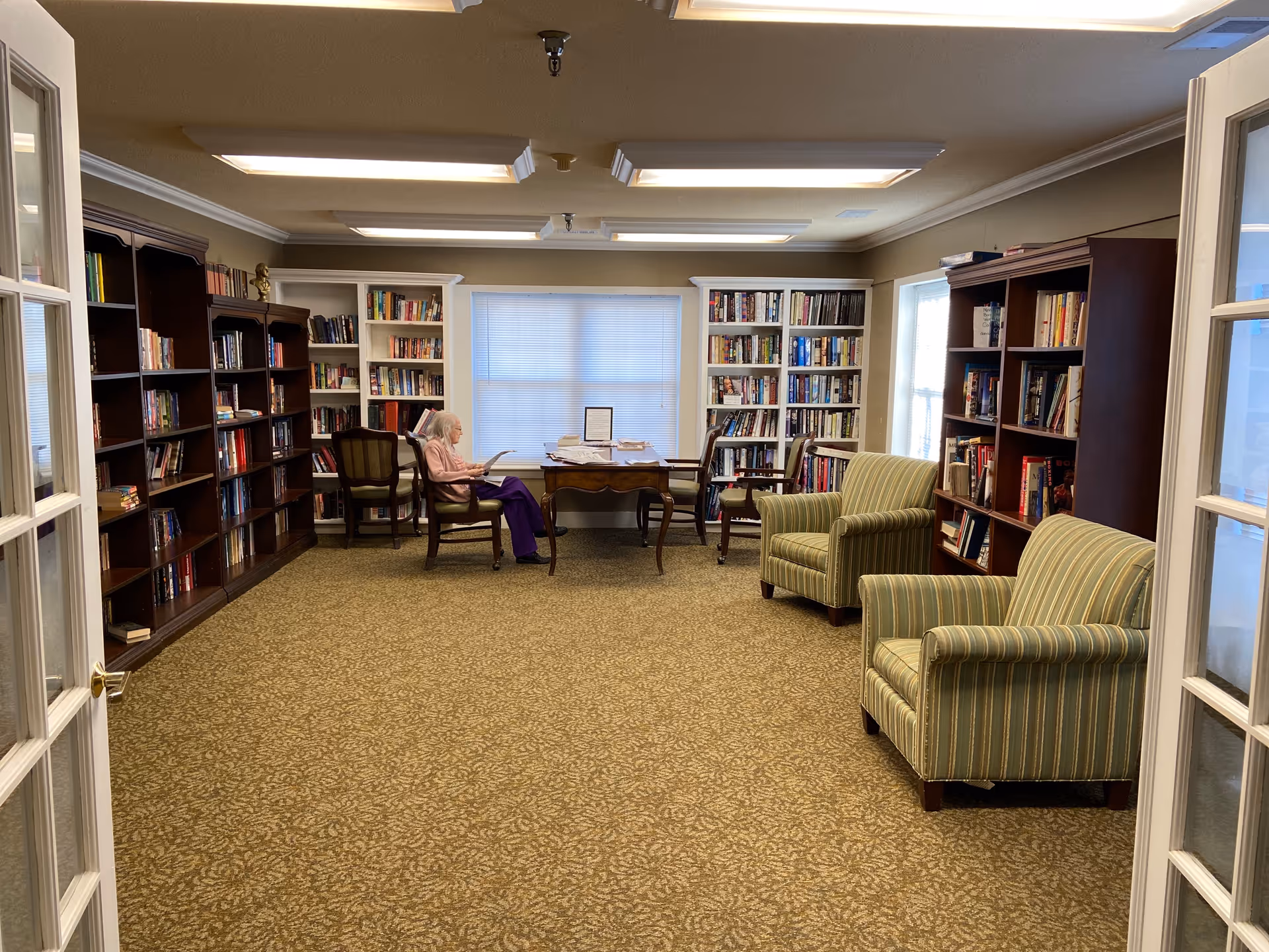 Carpeted community library room with bookshelves along the walls, upholstered armchairs, and a person seated at a table reading.