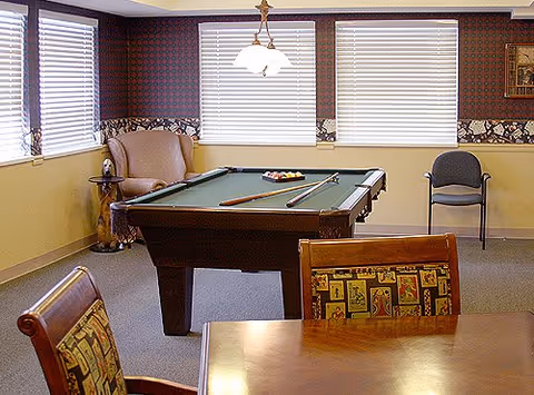 Interior recreation room with a pool table, cues, seating and a wooden table near windows with blinds.