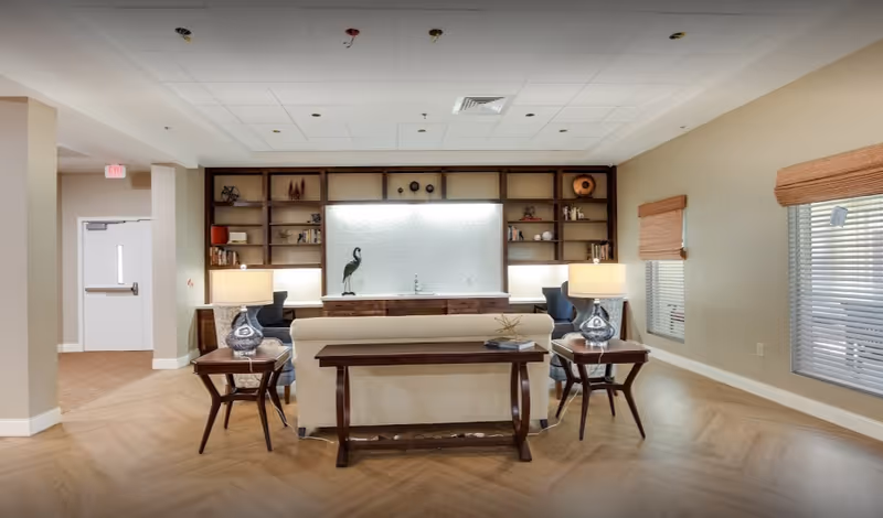A well-lit living room area in an assisted living facility featuring a beige sofa with a wooden console table behind it, two side tables with matching lamps, two blue armchairs, and a built-in wooden shelving unit with decorative items and books. The room has light-colored walls, wooden flooring, and windows with woven blinds.