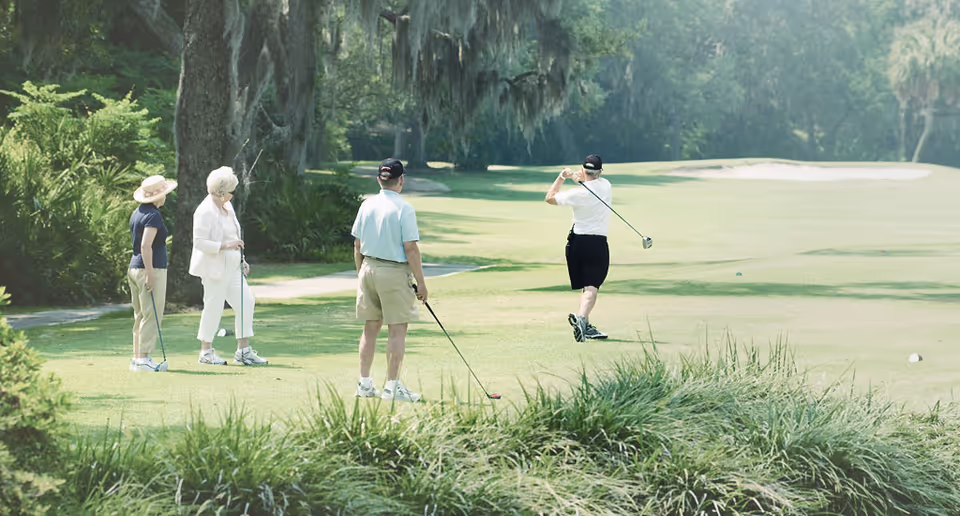 Four elderly people playing golf on a lush green golf course surrounded by trees and bushes. One person is in the middle of a golf swing while the others watch.