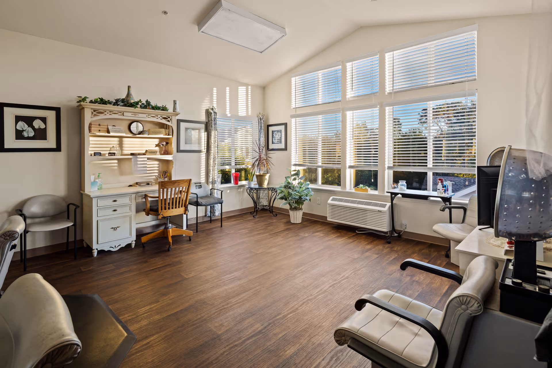 A bright room with large windows covered by white blinds letting in natural light. The room has wooden flooring and contains several chairs, a white desk with a hutch, a small table with plants, and a hair dryer chair, suggesting it is a salon or personal care area within a senior living community.