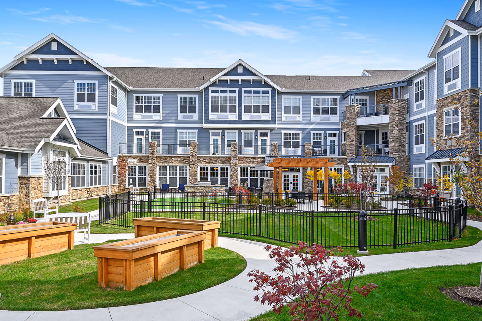 Courtyard with raised wooden planters and seating in front of a multi-story blue-sided senior living building with stone accents.